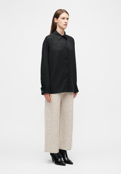 Γυναίκα που στέκεται φορώντας μια φαρδιά μαύρη πουκαμίσα με κουμπιά, φαρδιά μπεζ πλεκτή παντελόνα και μαύρα μποτάκια με τακούνι σε λευκό φόντο.
