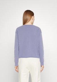 Femme aux longs cheveux bruns portant un haut à manches longues lavande et un pantalon blanc, vue de dos sur un fond uni.