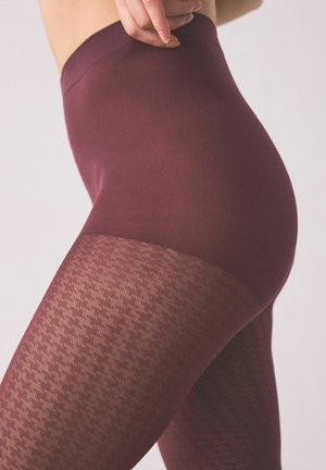 Nahaufnahme einer Person, die hoch taillierte, bordeauxrote gemusterte Strumpfhosen trägt, wobei eine Hand den Bund justiert.