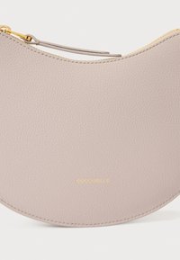 Lys beige skinnclutch med halvmåneform, gylden glidelås og preget logo. Strukturert overflate med glatt finish.