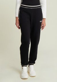 Pantalones de chándal negros en material suave con un corte ajustado, con rayas blancas a los lados y un logo en el muslo izquierdo. Un par de zapatillas blancas visibles.