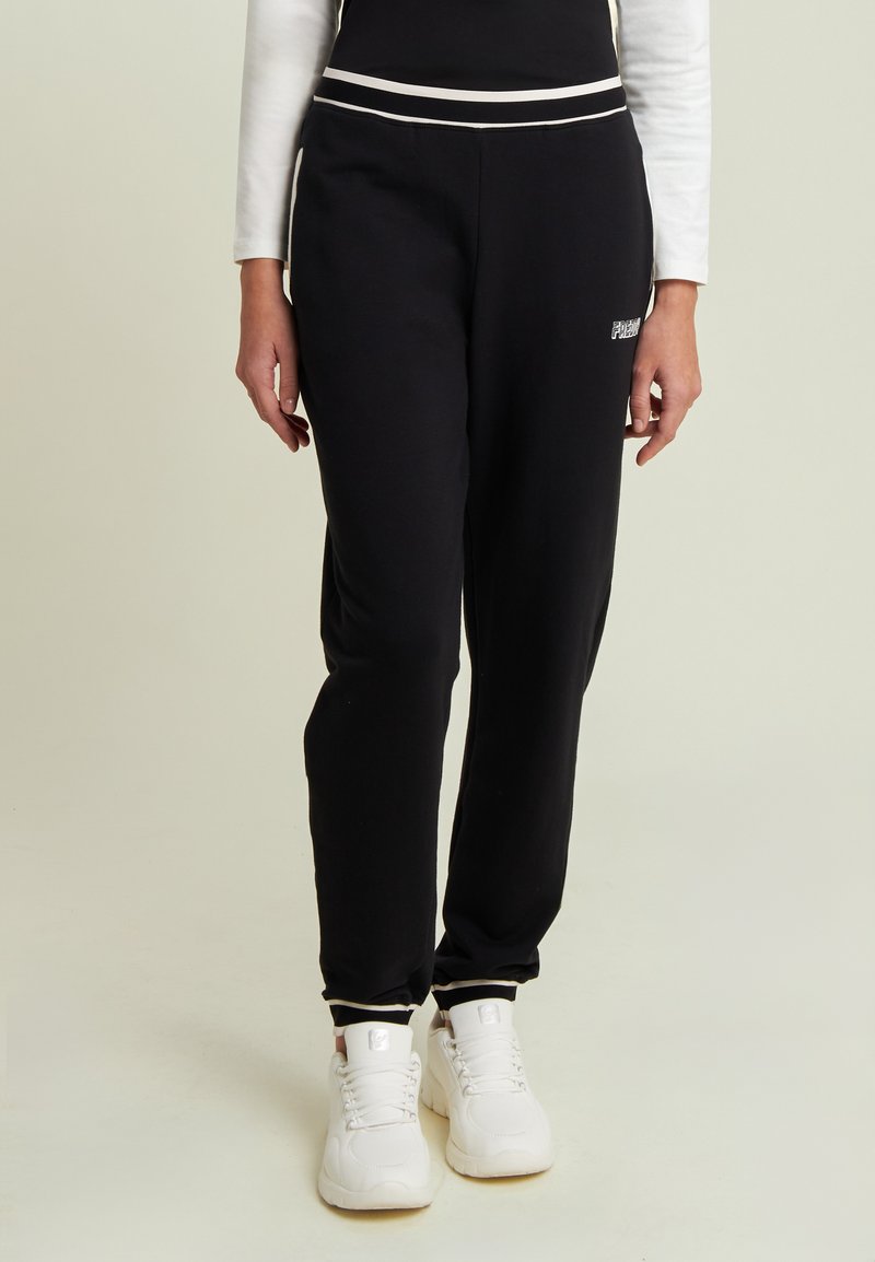 Pantalones de chándal negros en material suave con un corte ajustado, con rayas blancas a los lados y un logo en el muslo izquierdo. Un par de zapatillas blancas visibles.