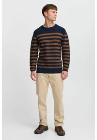 Pull rayé bleu marine et marron avec un col rond, associé à un pantalon cargo beige et des chaussures foncées. Couleurs unies et rayures horizontales.