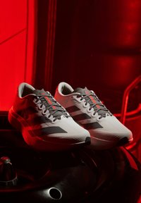 Weiße und graue Sportschuhe mit schwarzen Streifen und roten Akzenten, auf einer dunklen Oberfläche mit roter Beleuchtung im Hintergrund platziert.