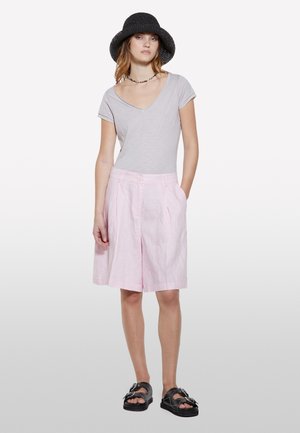 Maglietta grigia chiara a V, pantaloni corti rosa in lino con pieghe, cappello intrecciato nero e sandali neri con un accento a fiocco.