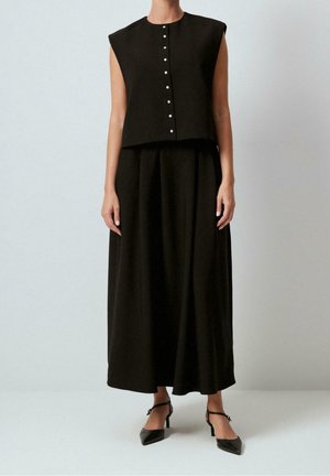 Kobieta ubraną w bezrękawnikowy czarny top zapinany na guziki, długą czarną spódnicę i czarne szpilki z ostrym czubkiem na jednolitym tle.