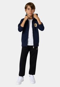 Granatowa bluza na suwak z logo na piersi, w połączeniu z czarnymi spodniami i białymi trampkami. Materiał wydaje się miękki, a fason casualowy.