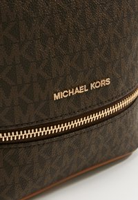 Sac Michael Kors marron avec motif MK embossé, logo en métal doré et détail de fermeture éclair dorée sur la poche avant.