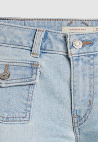 Détail d'un jean en denim bleu clair montrant la ceinture, passant de ceinture, bouton en métal, petite poche avant avec rabat, et étiquette indiquant "SUPERLOW BOOT".