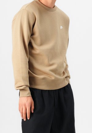 Mężczyzna w beżowej, długorękawowej bluzie Nike i czarnych plisowanych spodniach na jednolitym białym tle.