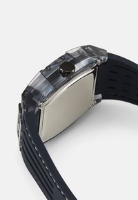 Transparente digitale Uhr aus Kunststoff mit einem navyblauen Silikonarmband, verfügt über ein rundes Gehäuse, einen einzelnen schwarzen Knopf und eine glatte silberne Rückseite.