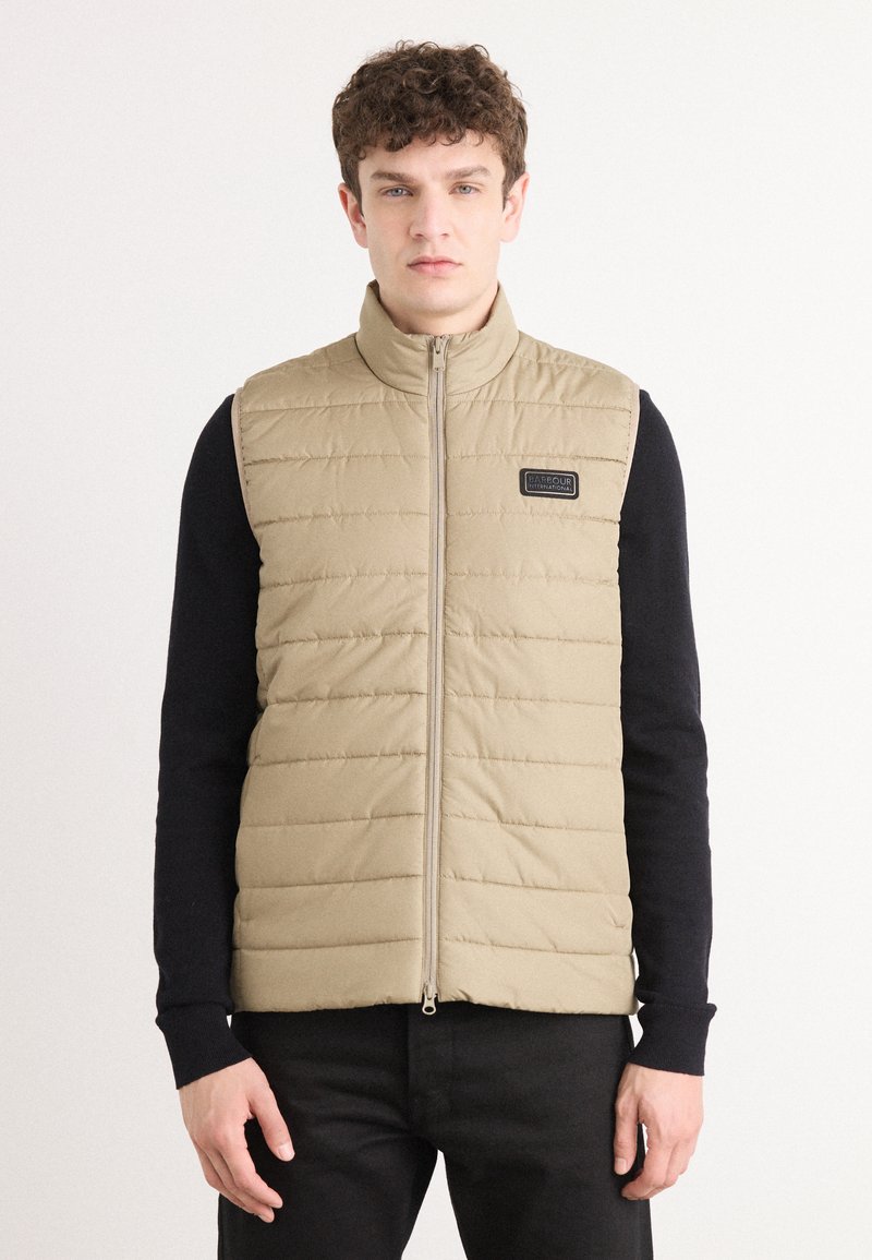 Μπεζ γούνα με ψηλό γιακά, φερμουάρ και λογότυπο. Φορεμένο πάνω από μαύρο πουκάμισο με μακριά μανίκια, που διαθέτει δομημένες ραφές.