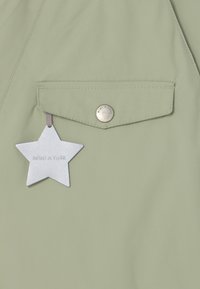 Lys grøn stof med en klaplomme sikret med en metallisk knap og et reflekterende sølvstjernetag mærket "MINI A TURE" hængende fra lommen.