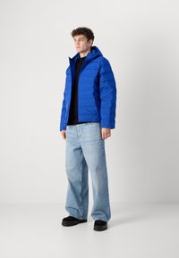 Chaqueta inflada azul con capucha, textura acolchada, cierre frontal de cremallera, combinada con pantalones vaqueros de pierna ancha azul claro y zapatos negros.
