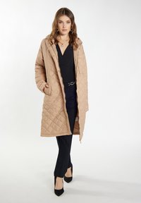 Gequilted beige lange jas met een capuchon, voorzien van goudkleurige accessoires en zijzakken, gecombineerd met zwarte broek en hakken.