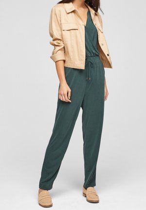 Kobieta w turkusowym kombinezonie z regulowanym pasem, beżowej krótkiej kurtce i pasujących beżowych mokasynach na płaskim obcasie, stojąca na białym tle.