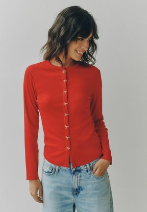 Donna con capelli mossi scuri che indossa una maglia rossa a maniche lunghe con piccoli bottoni a fiocco e jeans azzurri, sorridente e guarda verso il basso.