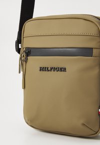 Geantă crossbody kaki realizată din material sintetic neted, având un fermoar negru, cusături albe și un logo "HILFIGER" reliefat.