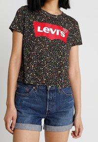 Camiseta negra de Levi's con estampado floral y mangas cortas, que presenta un gran logo rojo. Combinada con pantalones cortos de mezclilla azul con dobladillos doblados.