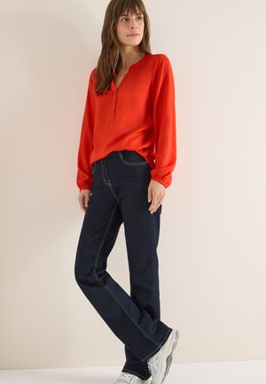Blusa rossa a maniche lunghe con scollatura henley e dettagli con bottoni, abbinata a jeans blu scuro con cuciture in contrasto e sneakers argento.