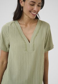 Blusa verde chiaro a v con maniche corte, tessuto testurizzato e bordo in pizzo decorativo lungo il colletto. Presenti sottili motivi verticali.