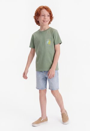 Uśmiechnięty chłopiec z rudymi włosami, ubrany w zielony t-shirt z nadrukiem, jasne jeansowe szorty i beżowe buty wsuwane, stoi na białym tle.