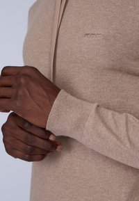 Pull en tricot beige avec une texture douce, à poignets ajustés et un détail de logo minimaliste. Gros plan sur des mains tenant le tissu.