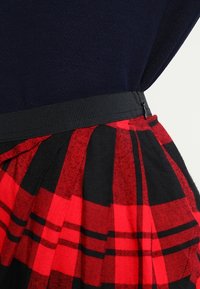 Musta ja punainen ruudullinen hame, jossa on kerrokselliset laskokset, mustalla vyötärönauhalla ja vetoketjukiinnityksellä. Pehmeä kangas, jossa on tekstuuri.