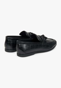 Zwarte instappers gemaakt van textuurleer, met een geweven patroon, platte zool en decoratieve kwastaccent aan de voorkant.