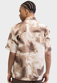 Kortærmet skjorte i creme med abstrakte brune mønstre, med et tekstureret stof og en standardkrave. Bagudvisning viser ingen bestemte hardware.