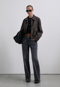 Woman wearing black sunglasses, cropped leopard print jacket, black crop top, gray flared jeans, holding large black bag, standing against white background.