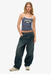 Top sin mangas azul con ribete blanco que presenta el texto "SUPERDRY BEACH CLUB", combinado con jeans de corte holgado de lavado medio en azul y zapatillas ligeras.