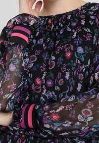 Blommig blus i genomskinlig svart tyg med lila, röda och blå mönster. Har elastiska ärmslut med rosa och svarta randiga detaljer.