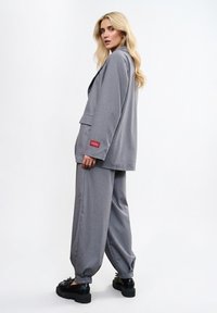 Sivý na mieru šitý blejzer a zodpovedajúce široké nohavice. Mäkká látka s hladkou textúrou. Čierne topánky na platforme. Detail loga na vrecku blejzera.