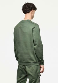 Hombre con cabello rizado que lleva una sudadera de color verde oliva y pantalones cargo a juego, de espaldas frente a un fondo claro y liso.