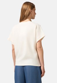 Top corto de color crema en tejido suave. Presenta un corte holgado, cuello redondo y puños doblados con un diseño liso en la parte de atrás.