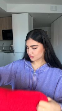 Lila Kapuzenpullover aus weichem Stoff, mit Reißverschluss und großen Taschen, gehalten von einer Person, im Vordergrund ein lebhaft rotes Kleidungsstück.