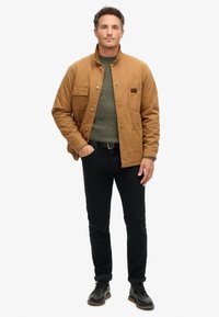 Chaqueta de trabajo marrón claro con botones a presión, dos bolsillos frontales y un parche de logo, combinada con un suéter verde y jeans negros con puños enrollados.