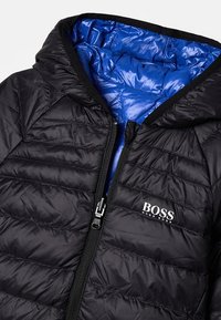 Chaqueta acolchada negra con forro interior azul, capucha y cremallera frontal. Presenta un logo blanco en el área del pecho izquierdo.