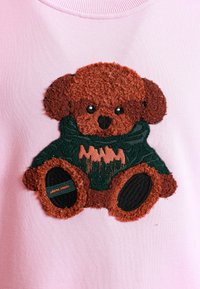 Sudadera rosa claro con un parche de osito de peluche texturizado en marrón, con ojos de botón negros y un suéter verde que presenta detalles en naranja.