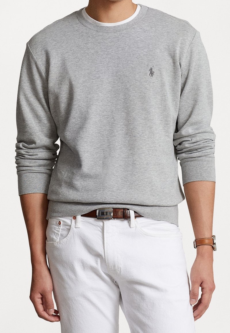Mann in einem grauen Langarm-Sweatshirt mit kleinem gesticktem Logo, weißen Hosen, braunem Leder-Gürtel und braunem Leder-Armbanduhr.