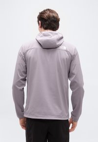 Άνδρας που φοράει ανο ιχτό γκρι μπουφάν με κουκούλα και το λογότυπο "The North Face" στην πάνω πλάτη, στέκεται μπροστά σε απλό λευκό φόντο.