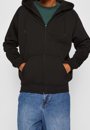 Person trägt einen schwarzen Zip-up-Hoodie über einem grünen Shirt mit Händen in den Vorderentaschen, kombiniert mit blauen Jeans.