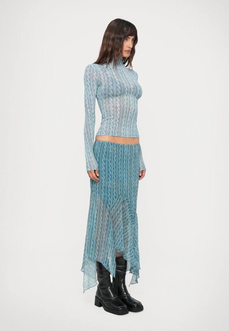 Woman wearing a blue patterned long-sleeve top and matching asymmetrical skirt with black heeled ankle boots, standing against a plain background.