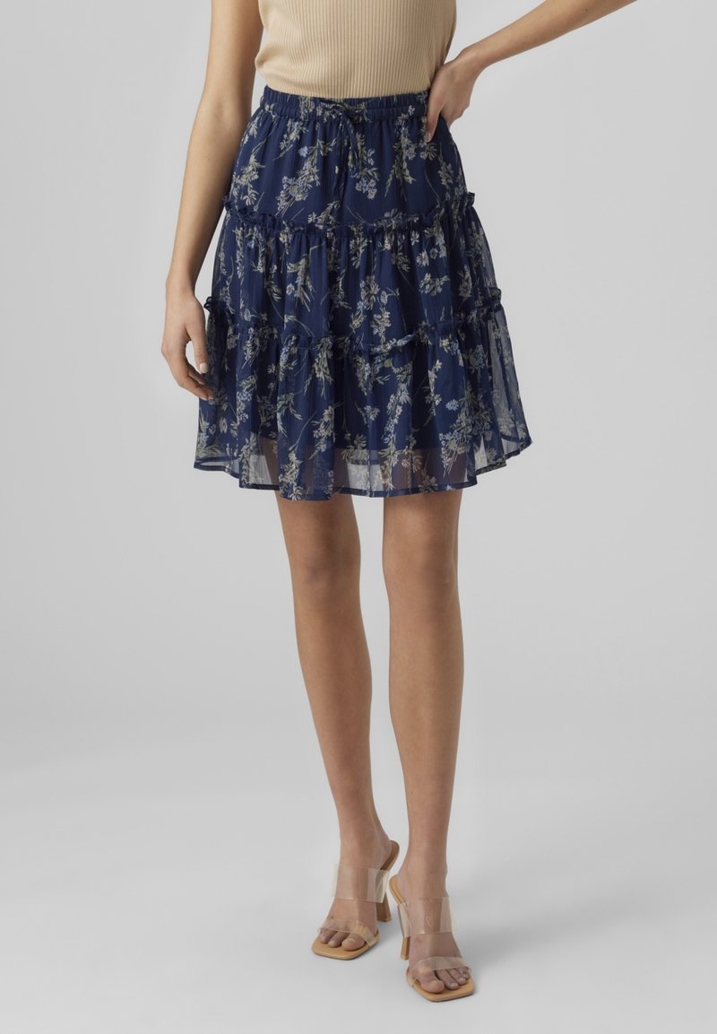 Femme portant une jupe à volants fleurie bleu marine avec taille élastique et superposition transparente, associée à un top beige sans manches et des sandales à talons transparentes.