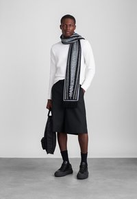 Fekete és szürke sál fráncos szélekkel, fehér pulóver fölött viselve, fekete sorttal párosítva. A modell egy fekete táskát tart, fekete tornacipőt visel.