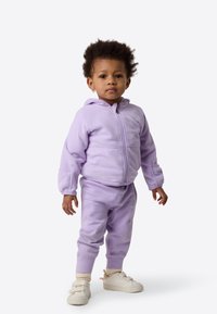 Lilla fleece hettegenser med glidelås og matchende bukser, med ribbestrikkede mansjetter og en liten logodetalj på benet, kombinert med hvite joggesko.