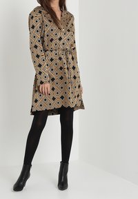 Braunes gemustertes Kleid mit geometrischen Designs, V-Ausschnitt, langen Ärmeln, tailliertem Bund und ausgestelltem Saum. Kombiniert mit schwarzen Knöchelstiefeln und Strumpfhosen.
