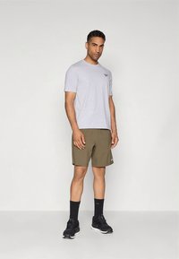 Szürke pamutpóló kis logóval, olívazöld rövidnadrággal párosítva. A modell fekete tornacipőt és bokazoknit visel, semleges háttér előtt áll.