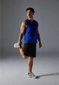 Conjunto atlético com uma camiseta azul sem mangas e calções pretos. A pessoa está a alongar-se, vestindo sapatilhas de corrida cinzas e brancas.